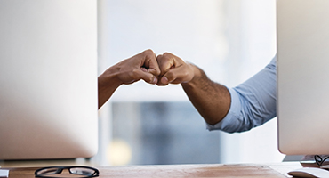 Two arms reaching out from behind computer monitors to bump fists